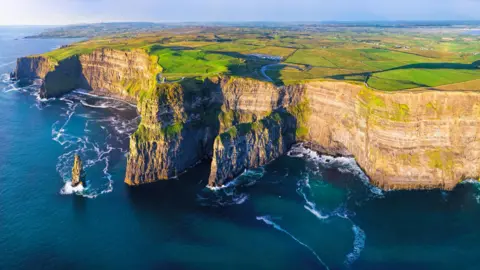 Getty Images The Cliffs of Moher viewed from above on a sunny day. The photo is a panoramic view of the faces of the cliffs, with a calm sea below. Above the cliffs, flat green fields stretch into the distance.