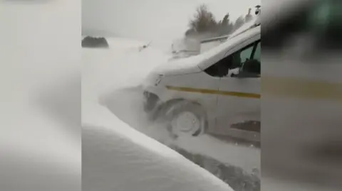 The front of a parked van is surrounded by a high wall of snow, which is still falling.