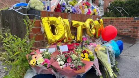 Bunches of flowers against a wall outside a house, with golden balloons spelling 'Dalton' above the flowers. Bunches of flowers on the top of the wall.