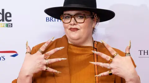 Jayde Adams in an orange blouse and jacket, wearing a black fedora hat. She is holding up her hands, which have long gold and silver nails and a chunky ring on each finger. She is wearing tortoiseshell glasses