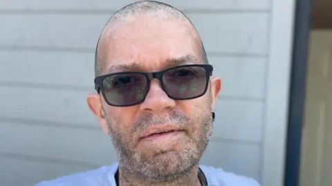 Stephen Huntley/BBC Jay House has very short grey hair and stubble. He is wearing black framed sunglasses, a blue T-shirt and has a black earring in one ear. He is standing in front of a wooden building.