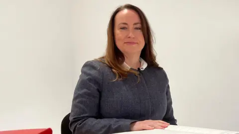 Claire Follain sat at a table with a large book. She is smiling at the camera and is wearing a grey jumper with a white collar. There are two books on the left. 