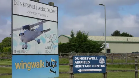 A sign which reads 'Dunkeswell Aerodrome' with a photo of a plane on it. There is also a sign saying 'Dunkeswell Airfield Heritage Centre'