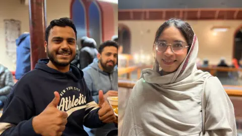 Mohammed is giving the thumbs up to the camera as he sits at a table with his food. He is wearing a navy blue hoodie with New York City Athletic written on it. He has short black hair push over to one side and a stubbly black beard. Amna smiles at the camera while wearing a white traditional Pakistani scarf. She has clear frame glasses on and behind her people are eating their meals.