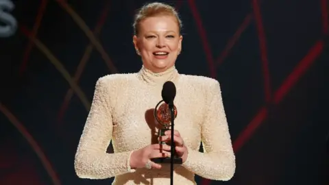 Reuters Sarah Snook accepts the Best Performance by an Actress in a Leading Role in a Play award for The Picture of Dorian Gray at the the 78th Annual Tony Awards in New York. she is wearing a cream high-neck dress with long sleeves.