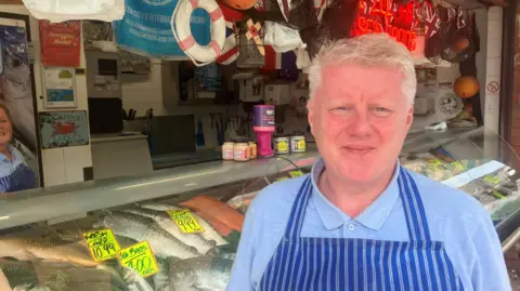Photograph of Mark Crossley in front of his fishmonger's stall at Oldham's Tommyfield Market. In the background are his fish.