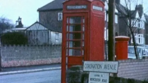 A 1970s photograph of a red telephone box with a postbox next to it. The image is slightly blurry and there are houses in the background.