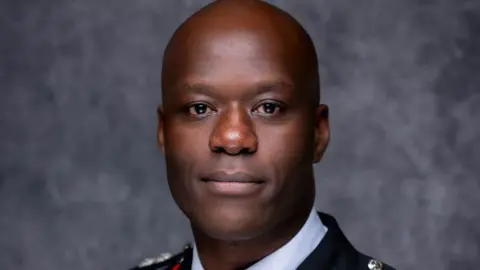 WMFRS A man with a shaved head and smiling face is wearing formal uniform and looking at the camera in a publicity shot. He is standing against a dappled grey background.