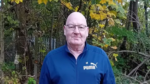 CancerResearchUK Martin Storey stood wearing a blue puma jacket against a fenced off area of woodland as he poses for a photograph. Martin is bald and wearing black rimmed glasses. 