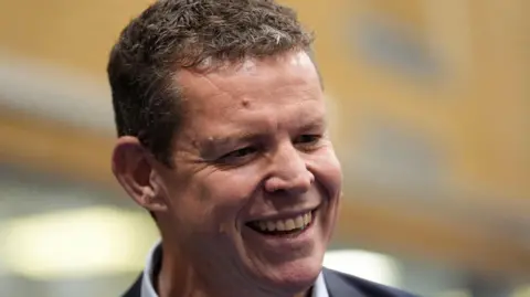 The image is a close-up of Plaid Cymru's leader Rhun ap Iorwerth. He is looking away from the camera and is smiling. 