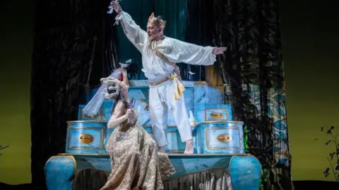 balletLORENT Geoff Hopson is in a white and gold costume looking down on the queen. He is wearing a gold crown and his arms are outstretched. The stage set is a huge representation of an ornate, blue and gold Regency style chest of drawers.