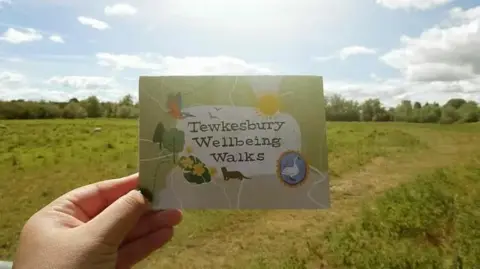 Gloucestershire Community Rail Partnership The back of the Tewkesbury Wellbeing Walks illustrated map, showing drawings of a swan, birds and trees. A woman’s left hand hold the map with countryside fields and trees in the distance.