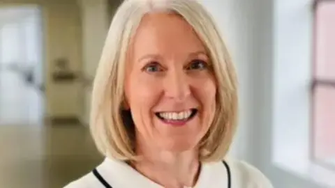 NHS Fife A woman wearing a light-coloured blouse with a black trim and a single dark button, standing indoors in a bright hallway with large windows.