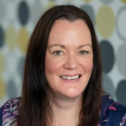 PA Media Kate Wallace, Victim Support Scotland. Woman with long dark hair, smiles looking into the camera wearing a navy blouse with pink and white details with a yellow and grey patterned wall out of focus behind her