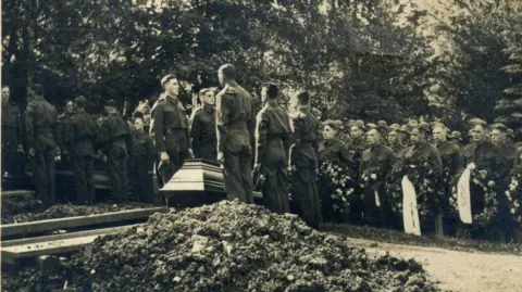 Libby MacRae A monochrome photograph from the 1940s which shows a burial ceremony in Berlin. In the foreground there is a pile of earth which has been removed from a freshly dug grave. Five soldiers in uniform can be seen standing to attention by a wooden casket. Further ranks of soldiers holding ceremonial wreaths stand in the background.
