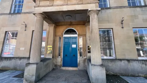The exterior blue double doors of the BLB Solicitors office in Trowbridge. It is a large sandstone building with covered porch area, supported by columns. here are lots of sash windows on either side of the arched entrance.