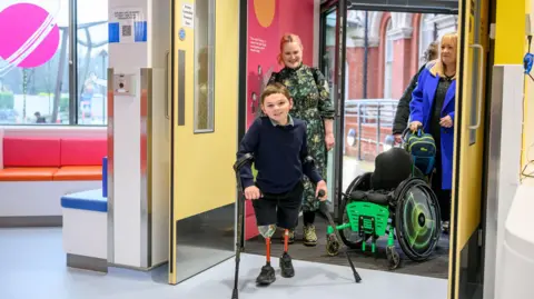 PA Media Tony Hudgell walks into a hopsital room on his prosthetic legs, with Paula entering behind him pushing his wheelchair.