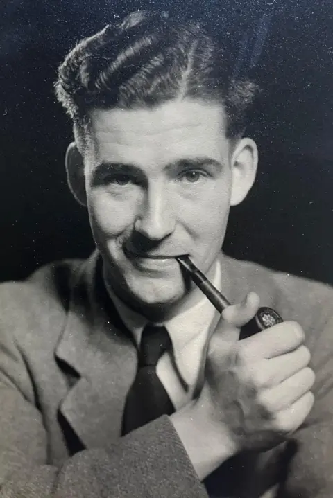 David Turner An old black-and-white photo of a man wearing a suit with a white shirt and a dark tie. He has dark curly hair, greased into a side parting, and is holding a pipe in his mouth with his hand on the bowl.