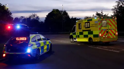 Getty Images A police car, with a closed sign on it, and an ambulance, on a road, at dusk. Trees are around the road. 