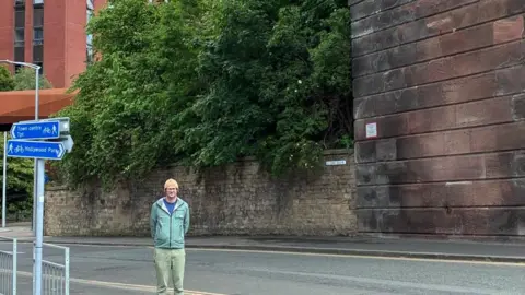 Kevin Bolton Archivist Kevin Bolton is wearing a turquoise rain jacket and green trousers. He stands under a large brick viaduct structure, not fully visible in shot. 