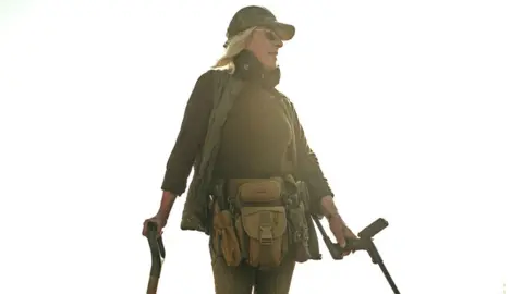 Nettie Edmondson Nettie Edmondson, looking to the left, with long blonde hair, sunglasses on, a cap, and green outfit, with a metal detector in one arm and a spade in the other. A large belt round her middle and a gilet. 