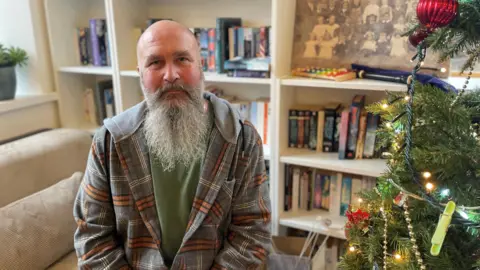A man with a bald head and a long, grey beard wearing a checked hoodie and a green jumper underneath. He is sitting next to a Christmas tree and in front of a book case.