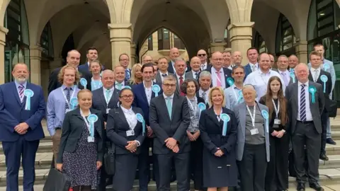 A group of Reform UK councillors are standing next to each other outside of a building. They are all wearing smart attire and blue rosettes. 