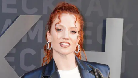 Jess Glynne looking at the camera. She is wearing a white t-shirt and a black leather jacket. She is wearing her hair in a ponytail and it is curly. The singer has a full face of make-up with long lashes and a nude lipstick shade on her lips.