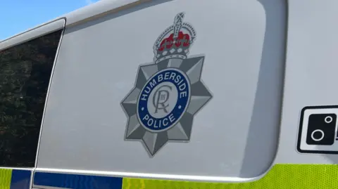 BBC A silver, blue and red Humberside Police logo on the side of a van. The logo features a crown, royal emblem and the words "Humberside Police". The van is white with yellow and blue graphics.