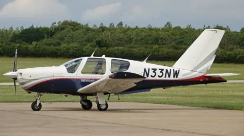 Side view on a runway of a Socata TB-20 Trinidad, a small white aircraft with a dark blue underside with N33NW written across it in dark blue