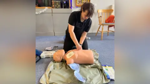 Contributed A woman is kneeling on the floor over a CPR dummy preparing to do a demonstration. She has dark hair with a fringe that is tied back. She is wearing a black T-shirt with black trousers and has her hands resting on the dummy's torso