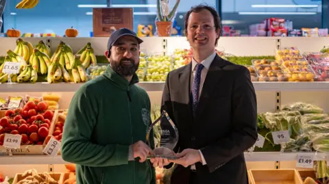 A fruit and veg stall holder holds the award with Alex Ross Shaw. The trader is wearing a green fleece and blue hat, while Ross Shaw is in a black suit.they are stood in front of a market stall selling fruit and vegetables, including bananas, tomatoes and grapes 