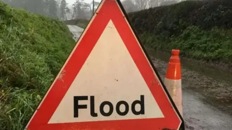 Axminister Fire Station  A triangular sign reads 'Flood', a cone stands behind it on a dirt track road with grassy hills at either side 