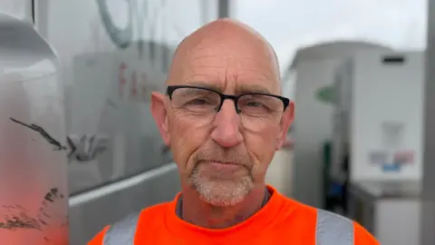 Steve Hubbard/BBC A head and shoulders image of John Mitchell. He is wearing an orange high-vis top and glasses. He is looking directly into the camera. 
