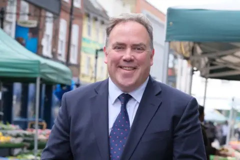 Croydon Council Jason Perry, wearing a navy suit and a spotty tie, smiling at an outdoor market