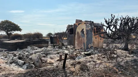 Remains of a small building surrounded by burnt grey remains on charred ground.