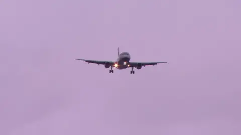 BBC A plane with lights on coming into land with a grey sky behind it