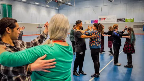 A group of people practice ballroom dancing in a sports hall with a blue floor. Everyone is lined up with a partner.