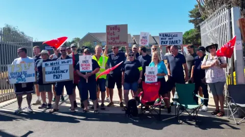 Strikers on a picket line outside the centre on a sunny day holding placards.