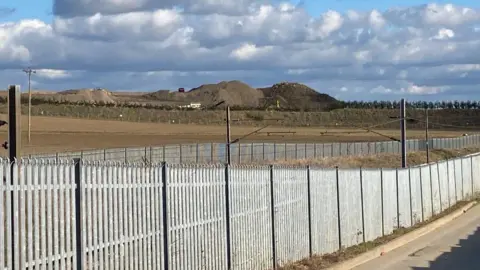 The waste recycling facility at Hacking Lane, South Elmsall. Large mounds of earth can be seen in the distance behind a metal fence.
