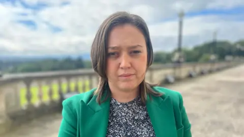 Deborah is wearing a green blazer and white and navy flowery high necked top underneath. 
She has short brown hair and is squinting at the camera with Stormont grounds in the background blurred in the shot. 