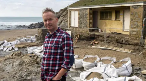 Mullion Surf Life-Saving Club representative Simon Mitchell stands on Polurrian Beach with the former lifeguard hut behind him. There are a lot of bags of building sand dotted in front of the building and metal fencing surrounding the hut. It is a cloudy day and Simon is wearing a red, white and blue checked shirt.