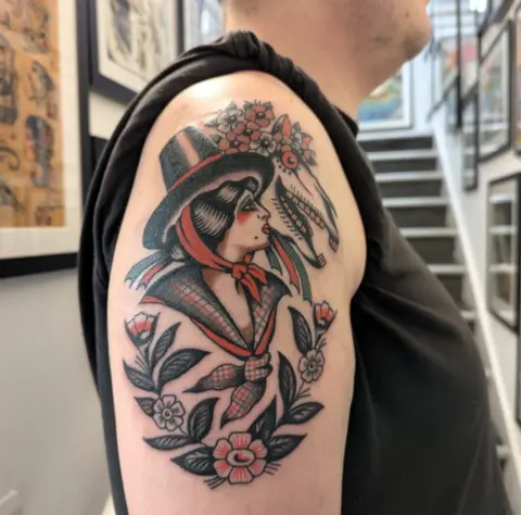 Chris Collins A traditional style tattoo on a man's upper arm. It features a woman in traditional Welsh clothing next to a Mari Lwyd - a horse skull decorated with flowers. 
