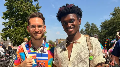 Harriet Morter/BBC Manny and Anthony standing next to each other in Norwich city centre outside The Forum. They are both looking directly at the camera and smiling and the sky is blue above them in the background. Manny is wearing a colourful abstract patterned shirt and apair of glasses. Next to him is Anthony who is wearing a cream short-sleeved shirt. He is wearing a pair of small hoop earrings and has a pair of sunglasses on his head.