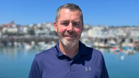Marc Leadbeater standing by the marina smiling to the camera. He has short grey hair and close-cropped beard and moustache. He's wearing a navy blue polo neck.