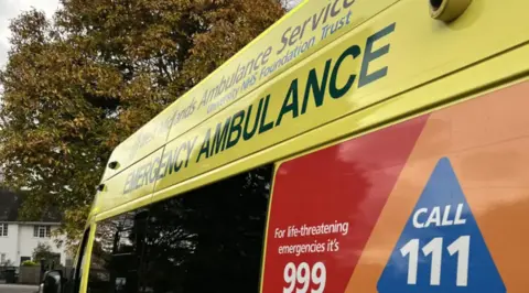A close up of the side of a yellow ambulance, with 999 and 111 numbers on the side and trees in the background