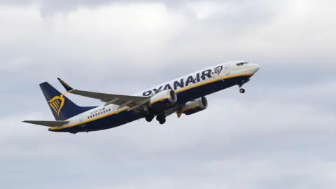 Getty Images A Ryanair plane flies against a background of mostly cloudy sky. 