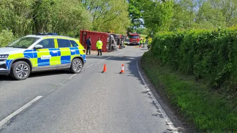West Mercia Police The same stretch of road as in the first image, taken from the other end. A police car is in the foreground with two red traffic cones, and the stricken lorry beyond. In the distance are fire crews and appliances.