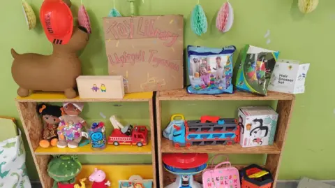 Toys displayed on a wooden shelving unit and cardboard sign saying Toy Library