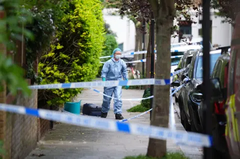 Police tape is tied across a street between trees as a forensic worker walks down it